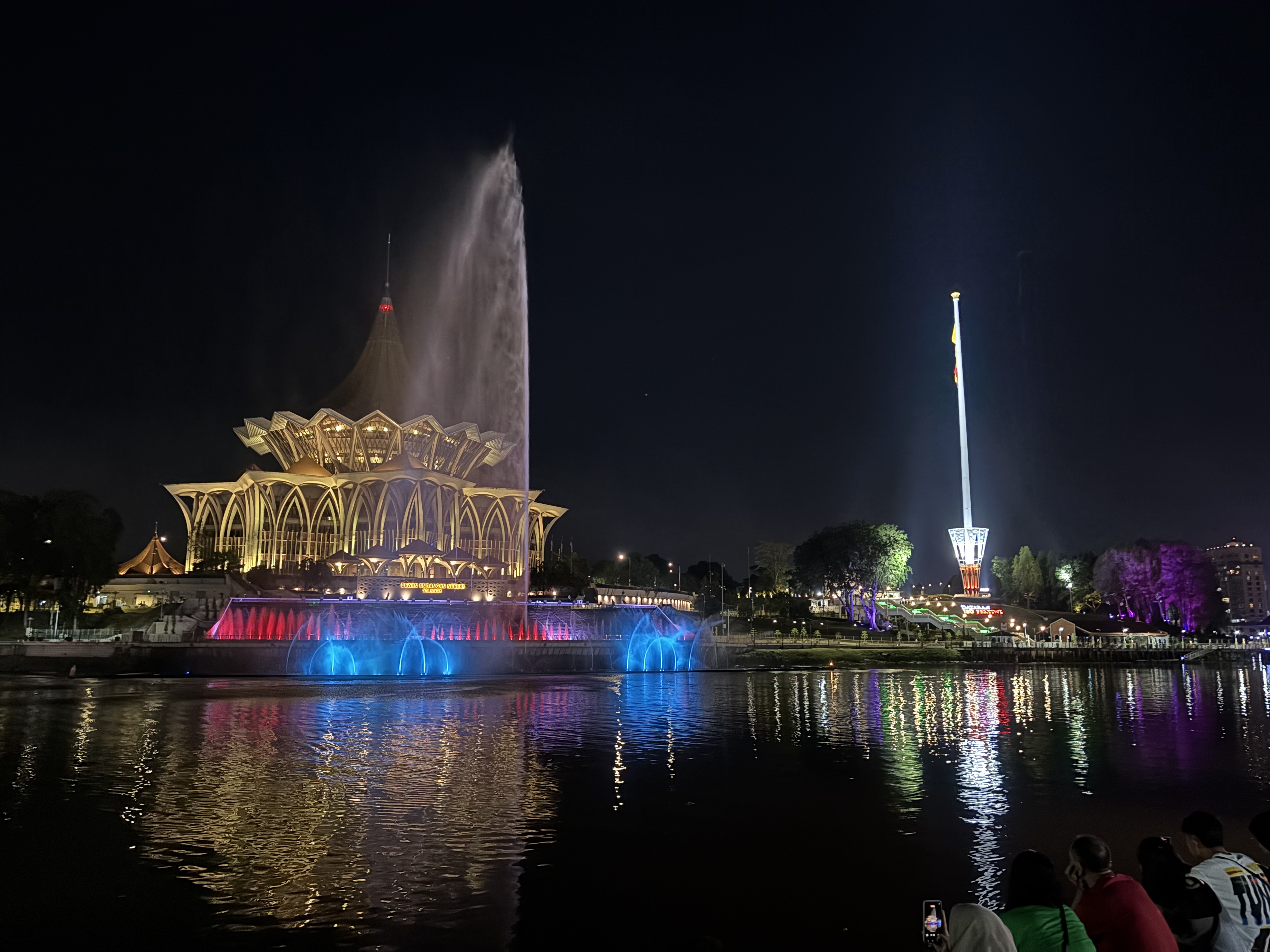 Fontana musicale del Darul Hana illuminata di notte a Kuching, con il Parlamento del Sarawak sullo sfondo e luci colorate riflesse nel fiume